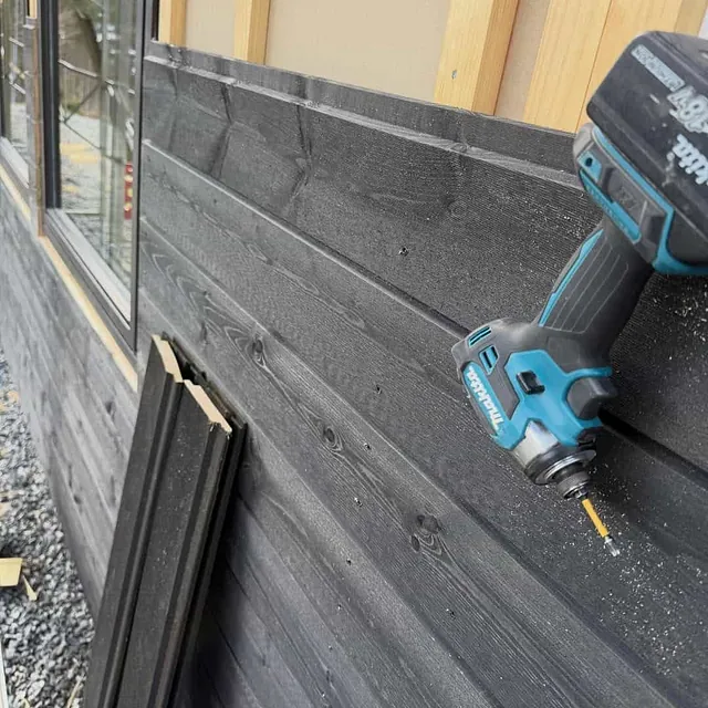 Nærbilde av mørk trekledning med synlige skruer og batteridrill under arbeid langs vegg.