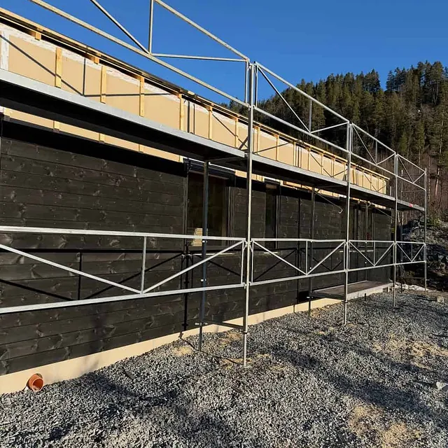 Fasade med ferdig montert kledning og stillas foran, omgitt av grus og skog i sollys.