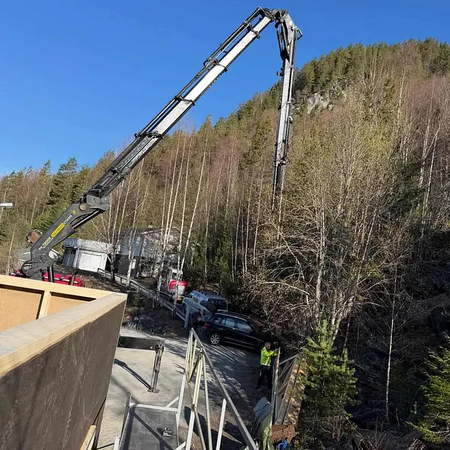 Lastebilkran løfter materiale på byggeplass ved skog og fjell, med stillas og bygg under oppføring.