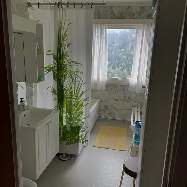 Bad med dusjforheng, plante og vindu med utsikt, innredning langs vegg i lyst rom.